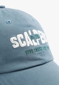 Lyseblå bomuldske med en struktureret design. Har hvid broderet tekst og grønne detaljer, herunder "SCALPERS" på forsiden.