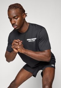 Černé sportovní tričko s krátkým rukávem s potiskem "ADIDAS RUNNING OD ROKU 1949" v bílé barvě, spárované s černými šortkami. Hladký, lehký materiál.