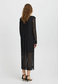 Rochie neagră cu mâneci lungi, cu dungi verticale și textură transparentă, având un tiv cu fante și asortată cu pantofi cu toc subțire negri.