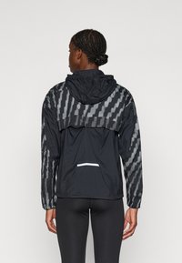 Giacca leggera nera con accenti riflettenti a motivo, cappuccio e tasca con zip nella parte posteriore. Maniche lunghe e vestibilità comoda.