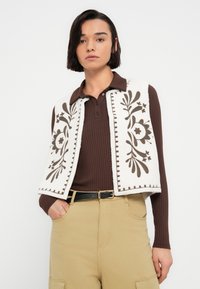 Veste en polaire blanche avec broderie florale marron et bordure ; portée sur un t-shirt à manches longues marron côtelé ; associée à un pantalon kaki et une ceinture noire.