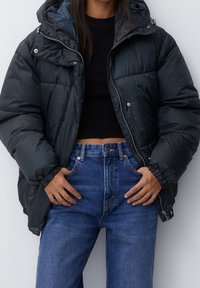 Sort puffermantel med hætte, der har en blank finish, lynlås og sidelommer, lagt over en sort top og blå højtaljede jeans.