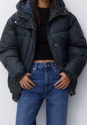 Giacca a vento nera con cappuccio, caratterizzata da una finitura lucida, chiusura a zip e tasche laterali, indossata sopra una maglietta nera e un paio di jeans blu a vita alta.