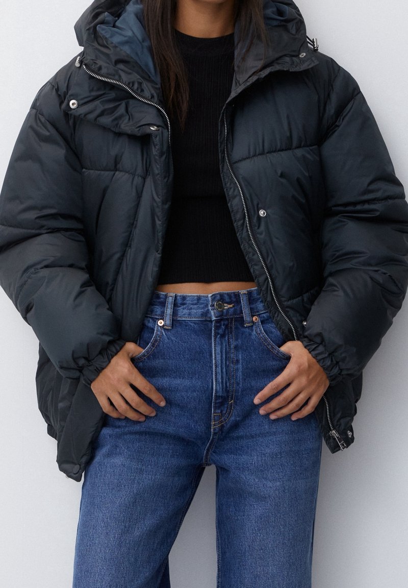 Sort puffermantel med hætte, der har en blank finish, lynlås og sidelommer, lagt over en sort top og blå højtaljede jeans.