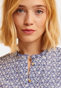 Blusa floreale blu con motivi bianchi a fiori, dettaglio con scollo arricciato e bottoni dorati. Tessuto morbido con maniche corte.