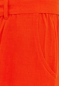 Shorts rossi in lino con vita piatta, tasche laterali e dettagli plissettati per una vestibilità rilassata. Texture liscia visibile in tutto il capo.