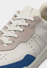 Κοντινή λήψη μπεζ και λευκού αθλητικού παπουτσιού με λευκά κορδόνια, διάτρητες πλαϊνές επιφάνειες και μπλε σουέντ λεπτομέρεια κοντά στη σόλα.