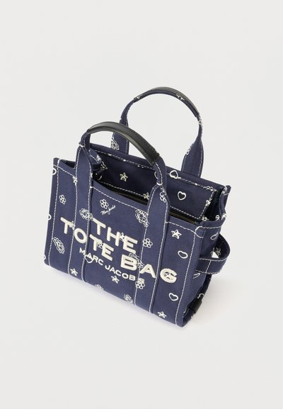 Geantă tote de culoare albastru marin de la Marc Jacobs, cu modele florale și stele albe, mânere din piele neagră și textul "THE TOTE BAG" pe față.