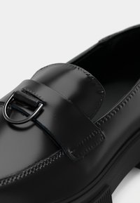 Mocassin en cuir noir à finition lisse, boucle métallique décorative et coutures détaillées le long des bords.