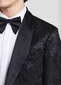 Giacca da smoking in velluto nero con motivo floreale intricatamente lavorato, risvolto in raso e tasca. Camicia bianca sotto, completata da un papillon nero.
