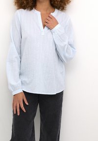 Ljusblå blus med långa ärmar, vertikala vita ränder, klassisk krage och strukturella detaljer. Kombinerad med vida svarta byxor.