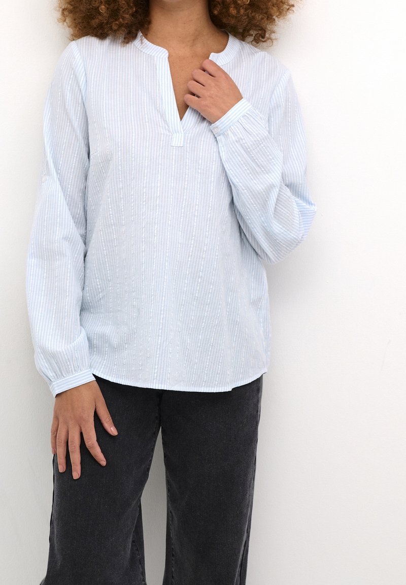 Ljusblå blus med långa ärmar, vertikala vita ränder, klassisk krage och strukturella detaljer. Kombinerad med vida svarta byxor.