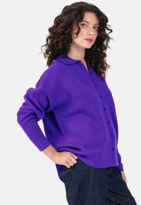 Cardigan violet tricoté avec col, fermeture boutonnée sur le devant et manches longues, fabriqué en matière douce ; porté avec un pantalon en denim foncé.