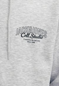 Pilkas džemperis su gobtuvu, pagamintas iš minkštos medžiagos; turi juodą siuvinėtą logotipą ir tekstą: "JACK & JONES Coll. Studio" su papildomu tekstu apačioje.
