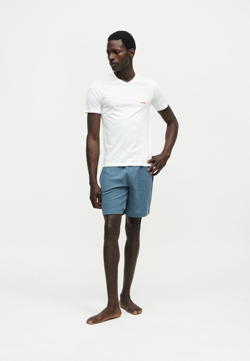Homme debout pieds nus, portant une chemise blanche à manches courtes avec un logo et un short bleu texturé au niveau des genoux, sur un fond uni.