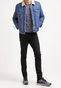 Giacca di denim con colletto in shearling, di colore blu. T-shirt grigia sotto, pantaloni neri e sneakers nere con motivo camouflage.