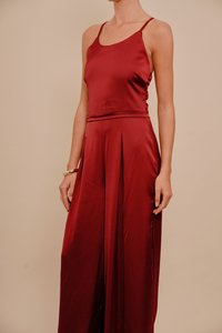 Kvinne iført en ærmeløs, burgunder satin jumpsuit med vide ben og et gullarmbånd på venstre håndledd, som står mot en beige bakgrunn.