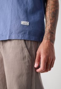 Camicia azzurra a maniche corte con etichetta tessuta, abbinata a pantaloni beige texture con tasche laterali. Visibili tatuaggi sulle mani.