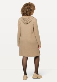 Abito beige in maglia con cappuccio, che arriva a metà coscia. Ha maniche lunghe e spacchi laterali all'orlo. Indossato con collant neri e scarpe beige.