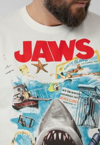 Weißes Baumwoll-T-Shirt mit einem lebendigen "JAWS"-Grafikdesign, das einen Hai, Taucher und Strandelemente in Blau-, Rot- und Gelbtönen zeigt.