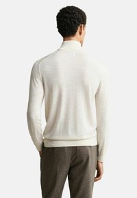 Mann mit lockigem Haar, der einen cremefarbenen langärmeligen Rollkragenpullover und braune strukturierte Hose trägt, mit dem Rücken zur Kamera gewandt.