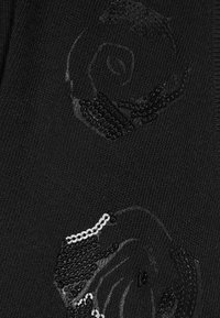 Tissu en tricot noir présentant deux motifs de rose ornés de sequins, ajoutant de la texture et un éclat subtil sur fond mat.