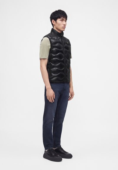 Hombre joven de pie vistiendo un chaleco negro acolchado, camisa beige de manga corta, vaqueros oscuros y zapatillas negras sobre un fondo blanco liso.