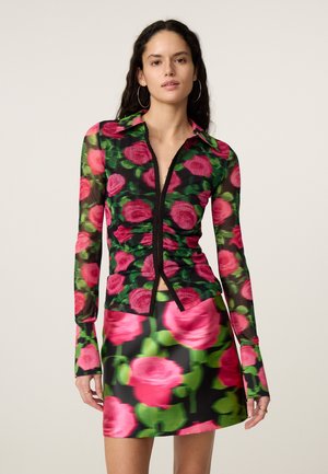 Vrouw die een nauwsluitende top met lange mouwen en een korte rok draagt, beide met levendige roze en groene rozenpatronen op een zwarte achtergrond.