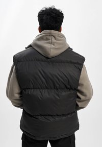 Kamizelka puchowa w kolorze czarnym, z pikowanym wzorem, noszona na szarej bluzie z kapturem, prezentując gładką fakturę i luźny krój.