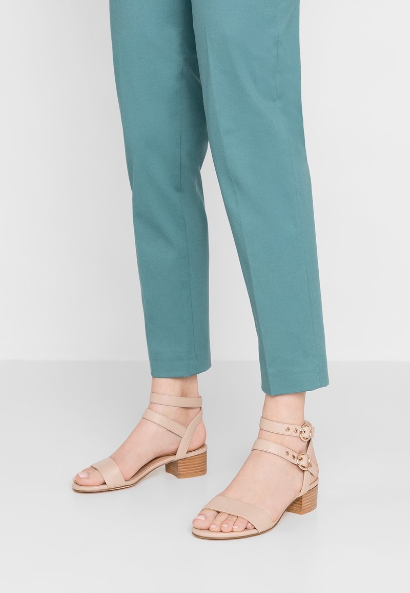 Sandales beige à lanières avec un petit talon carré, présentant des accents marron clair et des boucles réglables, portées avec un pantalon teal.