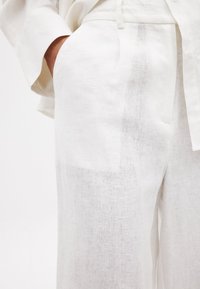 Baltas lina bikses ar brīvu piegriezumu, ar sānu kabatām un smalku tekstūru. Jostasvieta ir nostiprināta ar sasienamu lentu un izskatās viegla un gaisīga.