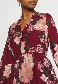 Vestido floral burdeos con mangas largas, que presenta flores en tonos rosa y crema, un escote en V y un diseño de cintura fruncida. Textura de tela suave.