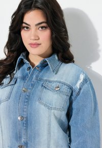 Vaaleansininen farkkutakki, jossa on kaksi rintataskua, hopeiset napit, kaulus ja kuluneet yksityiskohdat hartioissa.