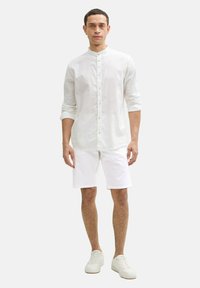 Weißes Leinenhemd mit Stehkragen, geknöpftem Vorderteil und langen Ärmeln, kombiniert mit weißen Shorts und weißen Sneakers, vor einem neutralen Hintergrund stehend.