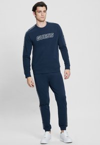 Completo felpa e pantaloni blu navy, con logo "GUESS" sul davanti, dettagli bianchi sulle maniche e polsini e vita a costine.