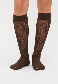 Calze al ginocchio di colore marrone, realizzate in pizzo con un motivo testurizzato e una fascia solida in cima. Il materiale sembra leggero e trasparente.