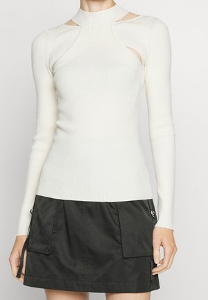 Mujer con una camiseta blanca de manga larga acanalada con aberturas en los hombros y una minifalda negra con grandes bolsillos delanteros.