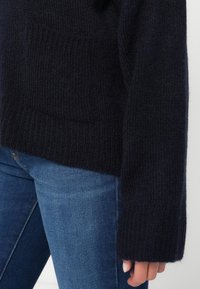 Granatowy sweter wykonany z dzianiny, z teksturowanym materiałem, ściągaczami przy mankietach oraz kieszenią z przodu. Połączony z niebieskimi dżinsami, tworzy swobodną kombinację outfitów.