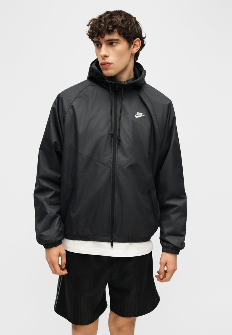 Giovane uomo che indossa una giacca a vento con cappuccio nera Nike e pantaloncini neri, che guarda verso sinistra su uno sfondo neutro.