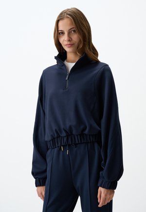 Navy half-zip pullover in soft fabric features elastic cuffs and hem, designed with a relaxed fit and subtle gold hardware accents.