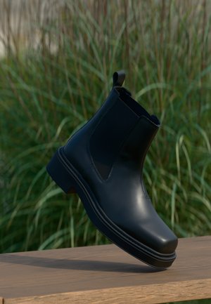 Μαύρο δερμάτινο μποτάκι Chelsea με ελαστικές πλευρές, στρογγυλή μύτη, χαμηλό τακούνι και θηλάκι στο πίσω μέρος, σε θολό πράσινο φόντο.