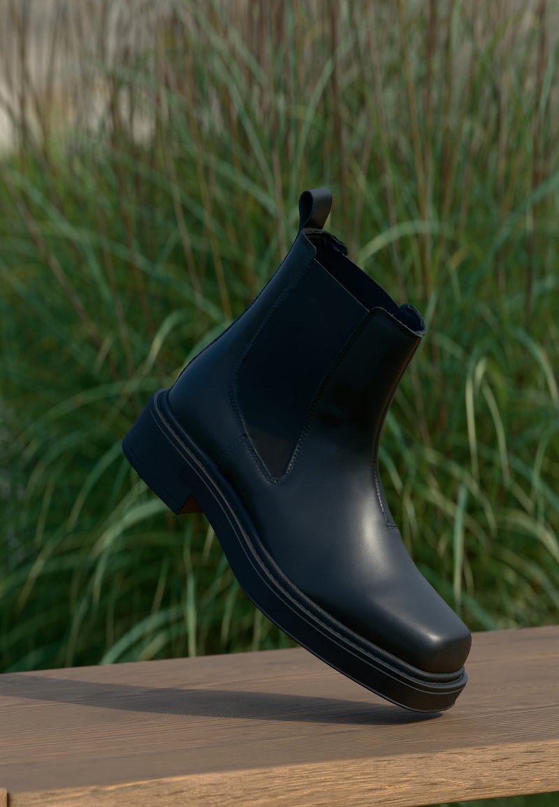 Musta nahkainen Chelsea-saapas, jossa on joustavat sivut, pyöreä kärki, matala korko ja vetotappi takana, sumentuneen vihreän taustan edessä.