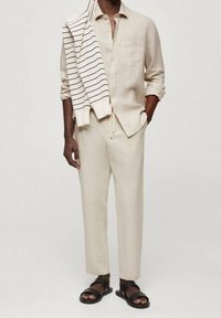 Jasnobeżowa lniana koszula i spodnie o luźnym kroju; kremowy sweter w paski zarzucony na ramiona; czarne sandały.