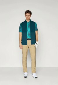 Costum de golf pentru bărbați care include o vestă teal, un polo de un albastru deschis, pantaloni bej și pantofi de golf albi; include un mănușă de golf pe mâna stângă.
