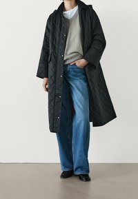 Zwarte doorgestikte lange jas met een kraag, gecombineerd met een grijze v-hals trui en wijde blauwe jeans. Zwarte schoenen maken de outfit compleet.