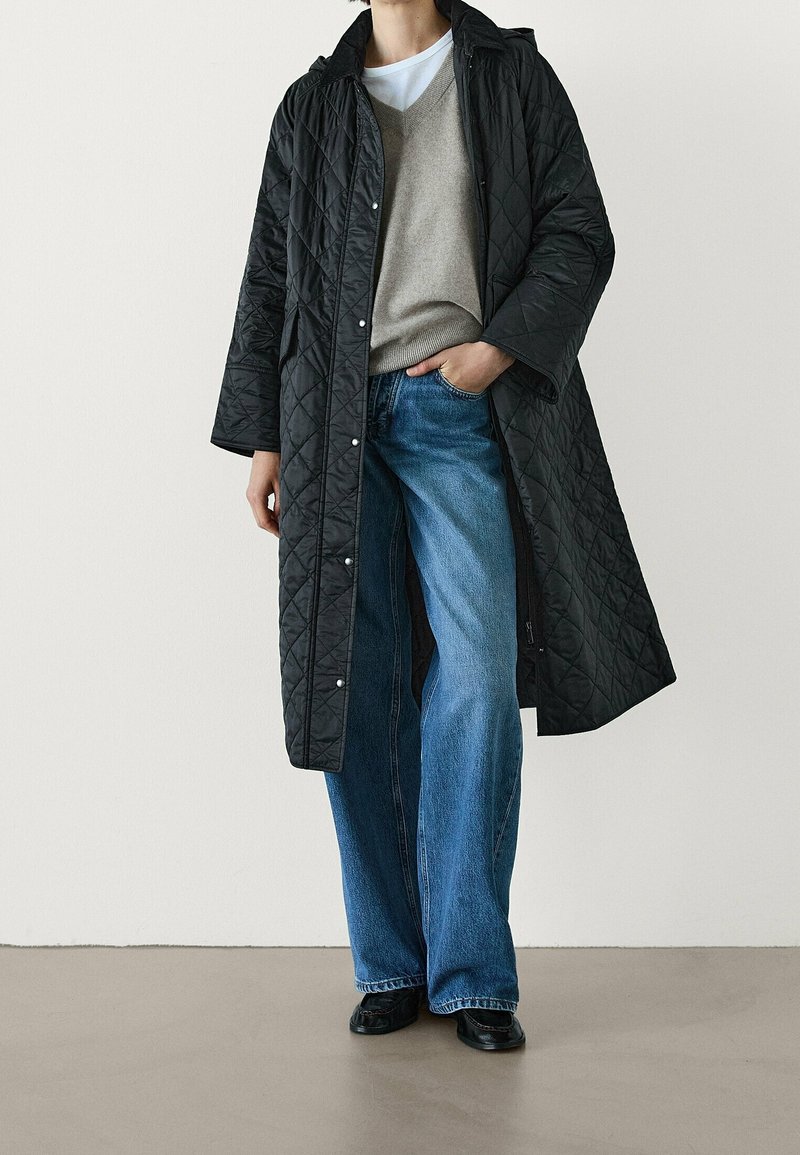 Zwarte doorgestikte lange jas met een kraag, gecombineerd met een grijze v-hals trui en wijde blauwe jeans. Zwarte schoenen maken de outfit compleet.