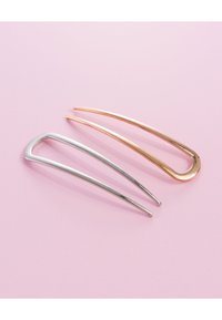 Kaksi tyylikästä metallista hiusneulaketta, yksi hopeinen ja yksi kultainen, asetettuna pehmeälle vaaleanpunaiselle taustalle, esittelee minimalistista muotoilua.