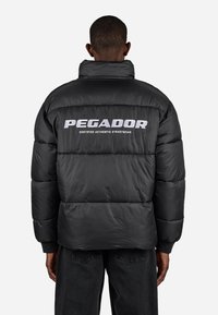 Mand iført en sort puffjakke med "PEGADOR Certified Authentic Streetwear" trykt i hvidt på ryggen, der står op ad en ensfarvet baggrund.