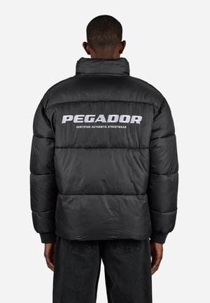 Mann, der eine schwarze Steppjacke trägt, auf der Rückseite mit "PEGADOR Certified Authentic Streetwear" in Weiß bedruckt, steht vor einem schlichten Hintergrund.
