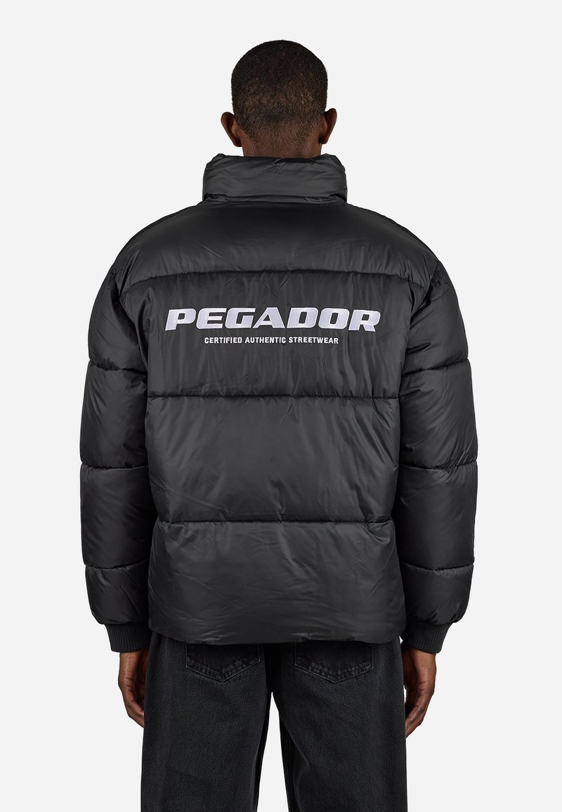 Mand iført en sort puffjakke med "PEGADOR Certified Authentic Streetwear" trykt i hvidt på ryggen, der står op ad en ensfarvet baggrund.